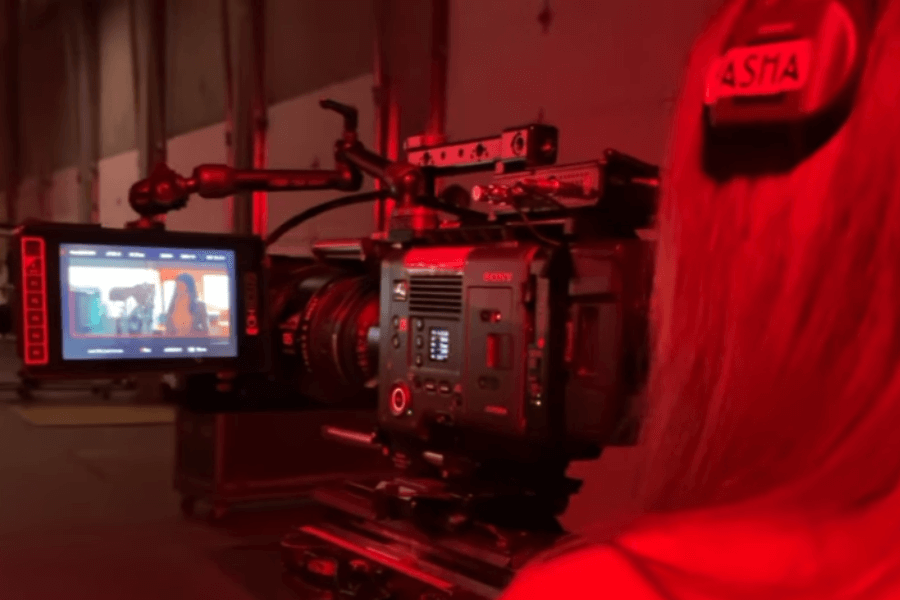 The image shows a close-up of a high-end cinema camera on set, capturing a scene with a red lighting atmosphere. The monitor attached to the camera displays a shot of a woman in the frame, likely from Woman of the Hour. A person, possibly Anna Kendrick, is seen from behind.