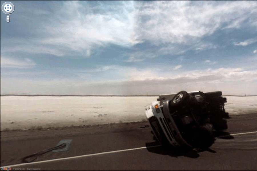 This image shows an overturned truck lying on its side on an empty road, with a vast, flat, and desolate landscape stretching out in the background under a partly cloudy sky. The flipped truck indicates a recent accident, possibly due to high winds, road conditions, or driver error, and the isolation of the area adds a sense of vulnerability to the image.