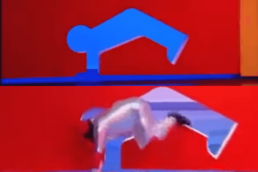 The image shows a contestant on the Japanese game show Hole in the Wall (also known internationally as Human Tetris), where players must contort their bodies to fit through cut-out shapes in a moving wall. 