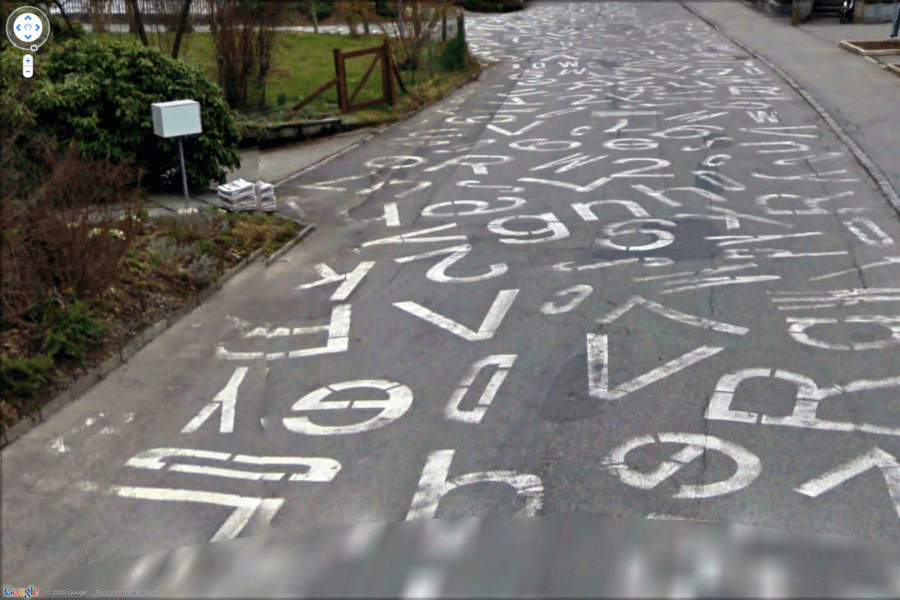 This image shows a road covered in a chaotic array of painted letters, numbers, and symbols, creating an abstract, almost artistic pattern across the surface. The markings appear random, with various fonts and sizes overlapping each other, transforming the street into an unconventional, visually striking scene. 