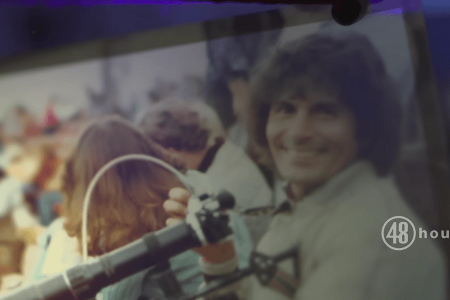 This image shows Rodney Alcala with curly hair smiling at the camera while holding what appears to be a camera or film equipment. 