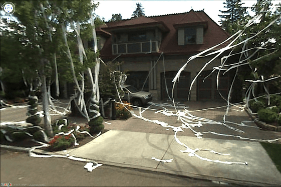 This image shows a house and yard that have been covered in toilet paper, with long strands draped over trees, bushes, and across the driveway. The scene resembles a classic prank, often associated with Halloween or mischievous celebrations. 