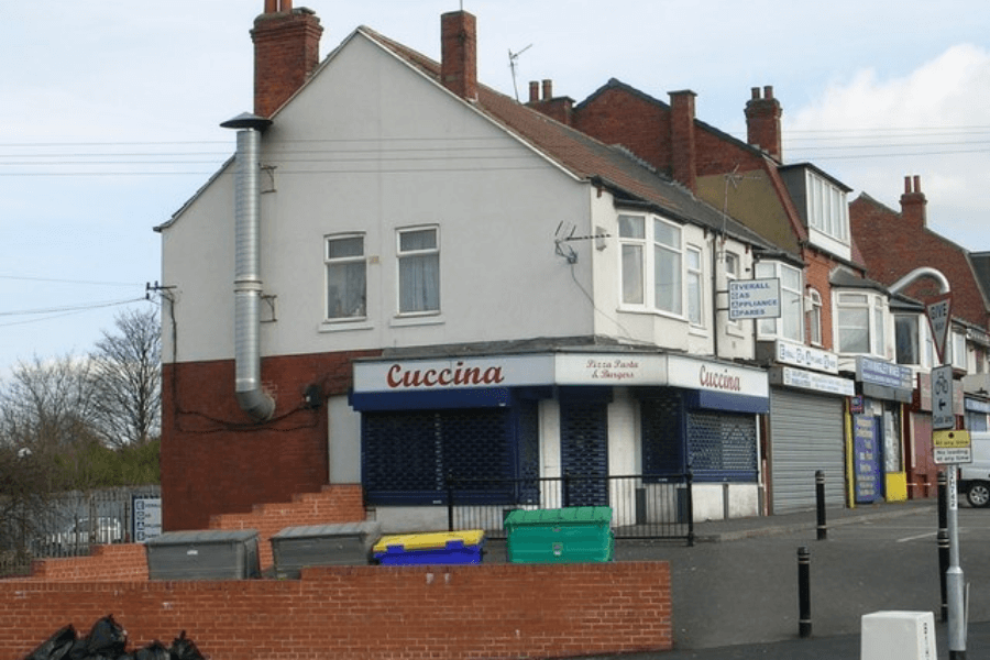 This image shows Cuccina, a modest corner restaurant with a brick facade and shuttered windows, suggesting a small business known for pizza and pasta, located in a quiet neighborhood with adjacent shops.