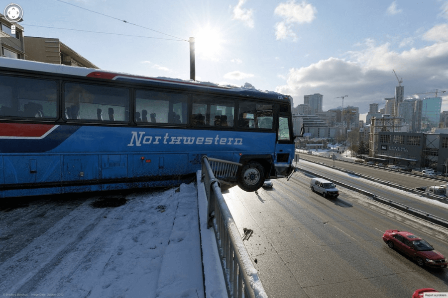 This image shows a blue bus labeled "Northwestern" hanging precariously over the edge of an elevated roadway or parking structure, with its front end suspended above a busy highway below. The snowy surroundings and clear sky suggest a winter setting, while a city skyline with tall buildings can be seen in the background. 