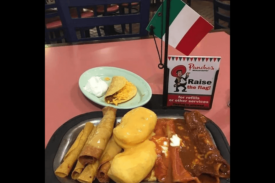 This image features a plate of classic Tex-Mex cuisine from Pancho's Mexican Buffet, including enchiladas, flautas, and sopapillas. The small table flag signifies their unique "Raise the Flag" service for refills or assistance.