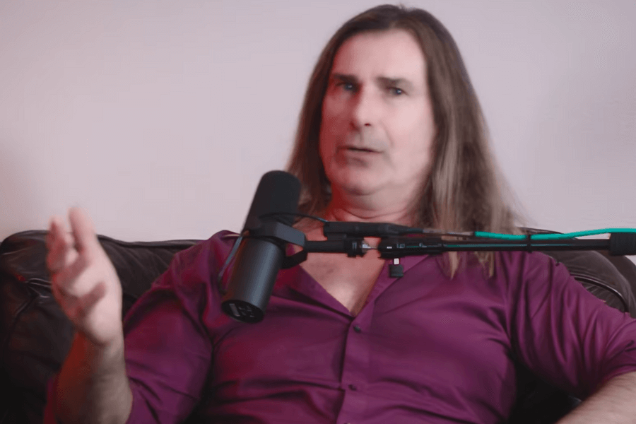 The photo shows a man with long hair wearing a deep purple shirt, sitting on a couch and speaking into a microphone during what appears to be a podcast or interview setting.