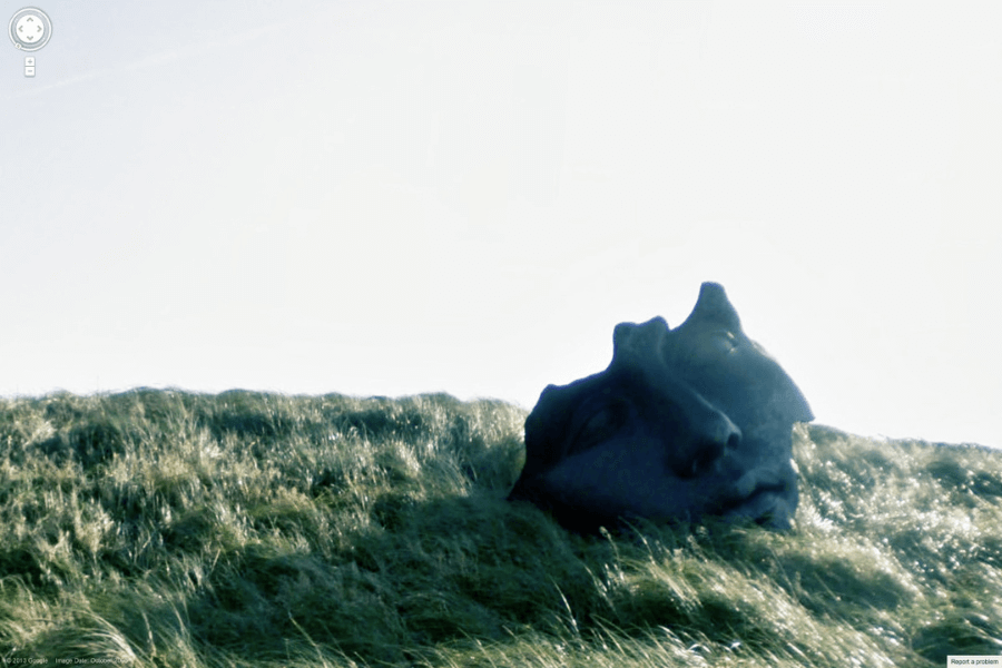 This image shows a large, sculpted face partially buried in a grassy hillside, giving the appearance of an ancient or surreal artifact abandoned in nature. The face has a serene expression, adding a sense of mystery and tranquility to the scene. 