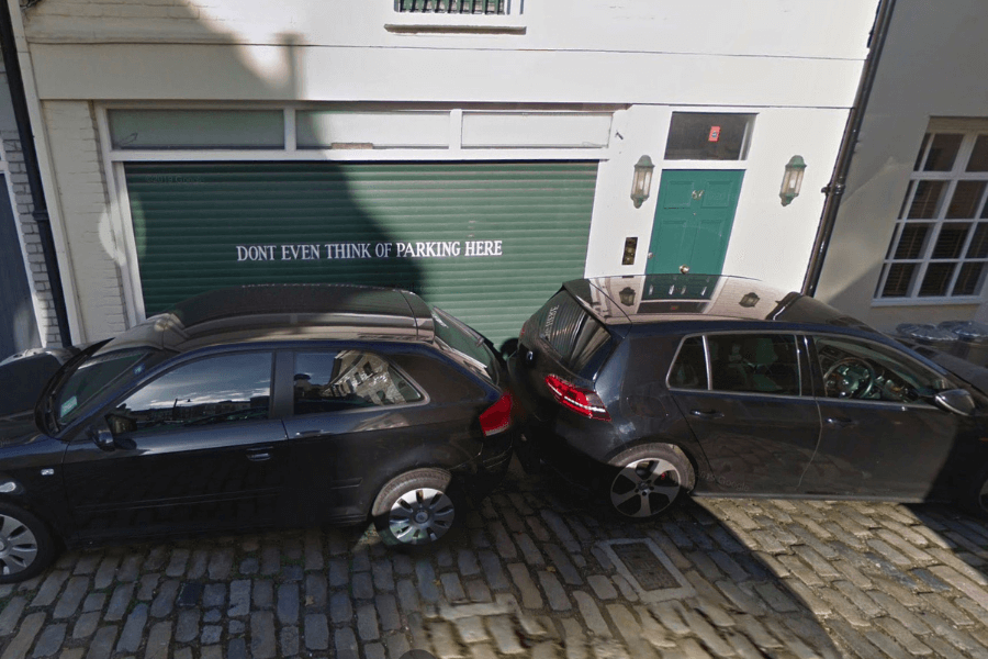 This image shows two cars parked very close together in front of a garage door with a bold message printed on it that reads, “DON'T EVEN THINK OF PARKING HERE.” Despite the clear warning, the two cars are positioned right in front of the door, seemingly defying the sign. 