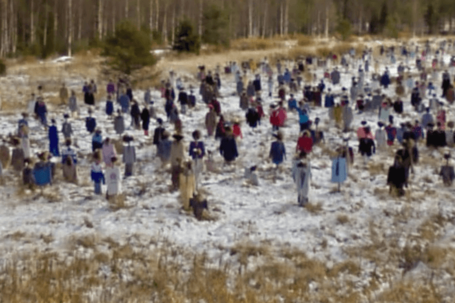 This image shows a field filled with numerous scarecrow-like figures dressed in various colorful clothes, hats, and garments. The figures are scattered across a snowy landscape, creating an eerie and surreal scene as they resemble a crowd of people frozen in place. 