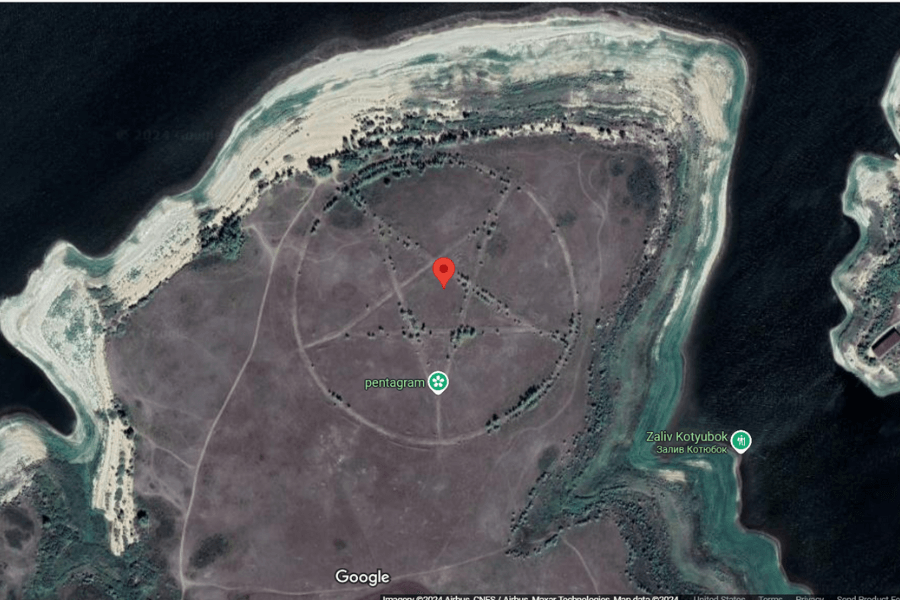 This satellite image shows a large pentagram symbol etched or formed in the landscape on a barren, isolated peninsula surrounded by water. The pentagram is clearly defined, with pathways or trees marking the lines and points of the star.