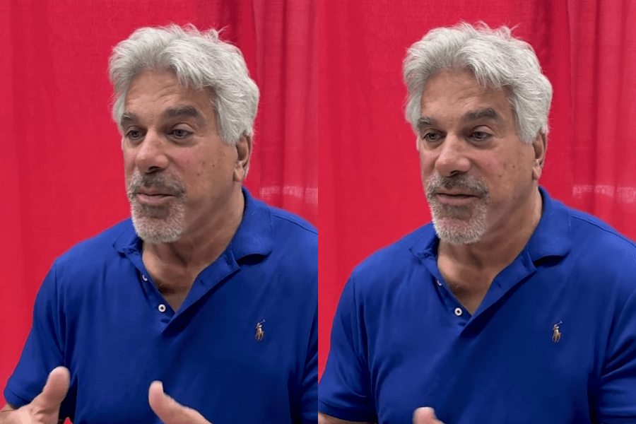 The photo shows a man with short gray hair and a beard wearing a blue polo shirt, gesturing with his hands as he appears to be speaking passionately, set against a red curtain backdrop.