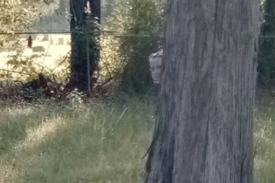 This image shows a partially obscured face peeking out from behind a tree in a wooded or grassy area. The person’s expression is unclear, adding an eerie and unsettling element to the scene, as they appear to be hiding or observing from the shadows. 