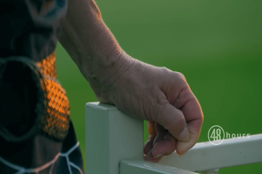 This image features a close-up shot of a hand gripping a white fence or railing, with the background blurred in a soft, green hue, indicating an outdoor setting. The hand appears to belong to an older person, with visible veins and wrinkles. 