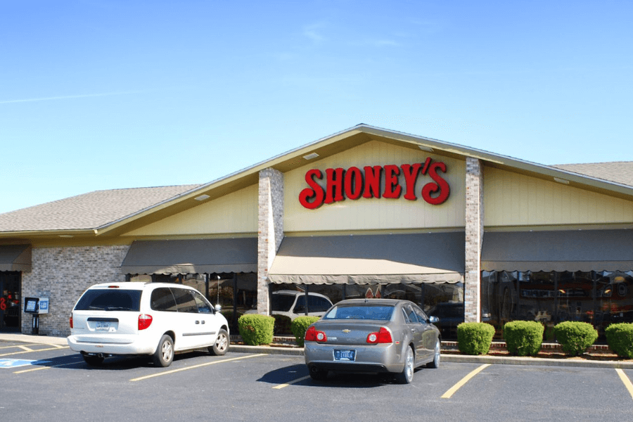 This image features a classic Shoney's restaurant, recognized by its signature red logo prominently displayed on the building. The exterior showcases a welcoming design typical of family-friendly dining establishments, with a spacious parking lot and neatly trimmed shrubs in front.