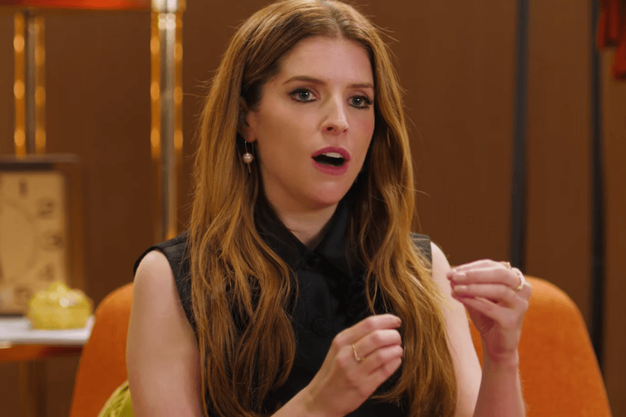 This image shows Anna Kendrick in a mid-conversation moment, gesturing with her hands and appearing engaged, possibly discussing a topic with intensity and passion, set against a warm, retro-styled background.