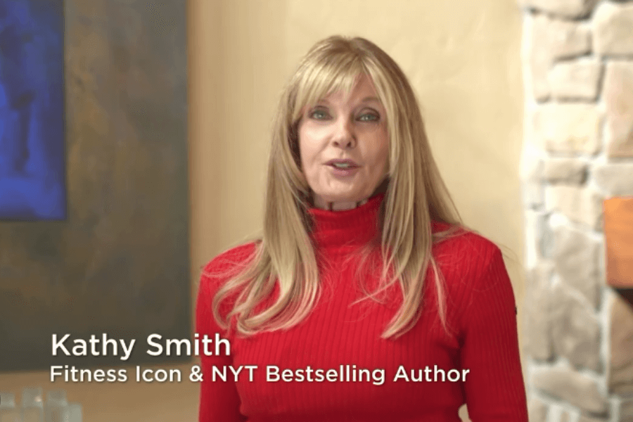 This image features Kathy Smith, the iconic fitness legend and NYT bestselling author, addressing viewers with her signature poise and charm. Dressed in a vibrant red sweater, she exudes warmth and confidence, reflecting her decades-long influence in the health and wellness industry. Behind her, a neutral, cozy setting complements her approachable yet professional demeanor, showcasing her evolution from an '80s fitness trailblazer to a respected thought leader today.