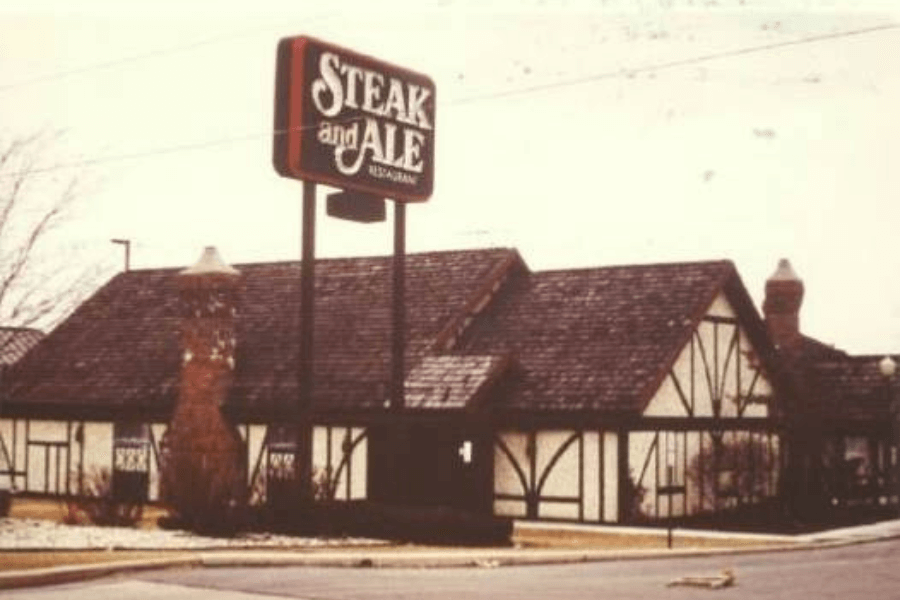 The image depicts a classic Steak and Ale restaurant with its Tudor-style architecture, featuring wooden beams and brick chimneys. A tall sign proudly displays the Steak and Ale logo, evoking a nostalgic dining experience.