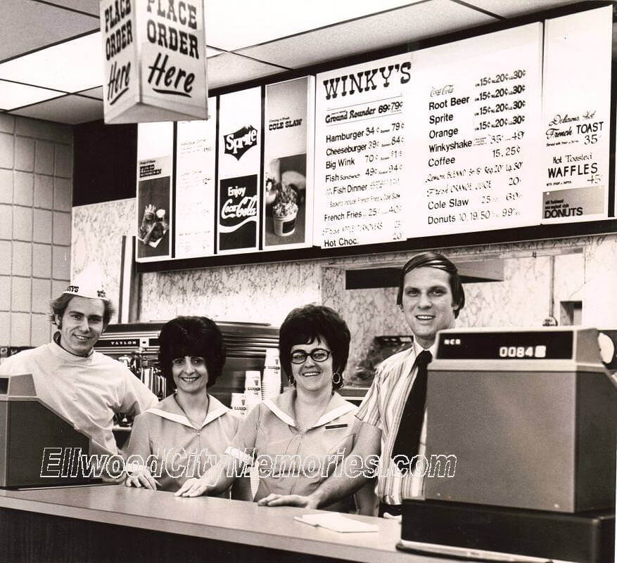 A vintage black-and-white photograph of Winky’s fast-food restaurant, featuring four smiling employees behind the counter. The menu board above lists classic items like hamburgers, cheeseburgers, fish sandwiches, and "Winkyshakes" with prices under a dollar. The scene includes an old-fashioned cash register and a hanging "Place Order Here" sign, capturing the nostalgic atmosphere of mid-century fast food.