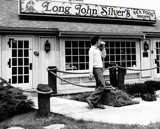 A black-and-white vintage photograph of a Long John Silver’s Seafood Shoppe featuring a classic nautical-themed exterior with wooden posts and rope railings. The restaurant’s sign, adorned with a pirate mascot, hangs above the entrance with large windows and lantern-style lighting fixtures. A young man in casual attire leans against a post while drinking from a cup, adding to the nostalgic feel of the scene.