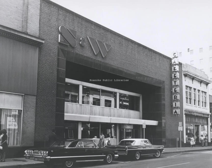 A vintage black-and-white photograph of an S&W Cafeteria building with a modern mid-century architectural design, featuring a sleek black entrance and large windows. The word "CAFETERIA" is vertically displayed in illuminated block letters on a sign to the right of the building. Classic cars from the 1960s are parked along the street, and pedestrians are seen walking by, adding to the historical atmosphere.