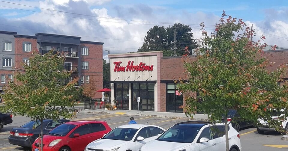 A Tim Hortons café with a modern brick exterior and large glass windows sits in a suburban shopping area. The restaurant's red logo is prominently displayed above the entrance, and an outdoor seating area with red umbrellas is visible to the side. Several cars are parked in the lot, and trees with autumn-colored leaves line the foreground.