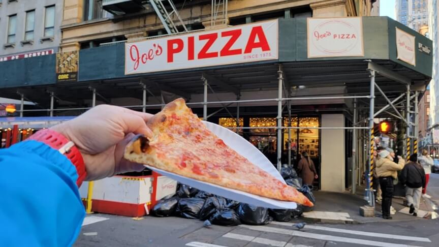 A person holds up a large, thin slice of cheese pizza on a white paper plate in front of Joe’s Pizza, a well-known pizzeria in New York City. The restaurant's red and white sign reads "Joe’s PIZZA Since 1975," and the storefront is partially covered by scaffolding. Pedestrians walk by in the background, adding to the bustling city atmosphere.