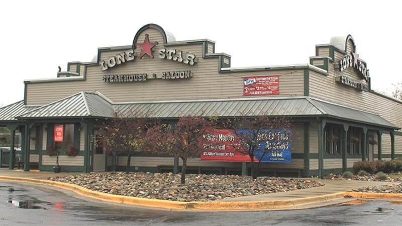 A Lone Star Steakhouse & Saloon restaurant with a rustic Western-themed exterior featuring beige siding, a green metal roof, and a large red star in its signage. Promotional banners advertising specials hang on the front, and a neon "OPEN" sign is visible near the entrance. The surrounding landscape includes decorative rocks, small trees, and a wet parking lot reflecting an overcast sky.