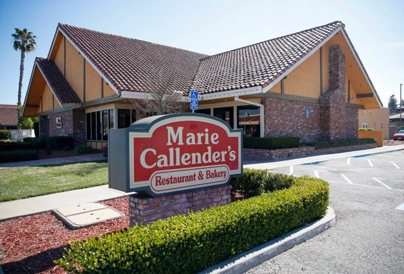 A Marie Callender's Restaurant & Bakery with a classic suburban design, featuring a steeply pitched tiled roof, brick and stucco exterior, and a large chimney. A red and white sign in the foreground displays the restaurant’s name, mounted on a short brick base surrounded by neatly trimmed hedges. The parking lot has handicap spaces near the entrance, and palm trees in the background suggest a warm climate.