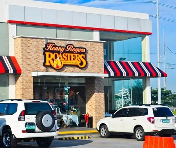 A Kenny Rogers Roasters restaurant with a modern glass-front design, a brick facade, and red, black, and white striped awnings. The illuminated sign displays the brand’s signature flame logo and the tagline “Deliciously Healthy.” Several white SUVs are parked in front, and the sky is bright and clear, suggesting a sunny day.