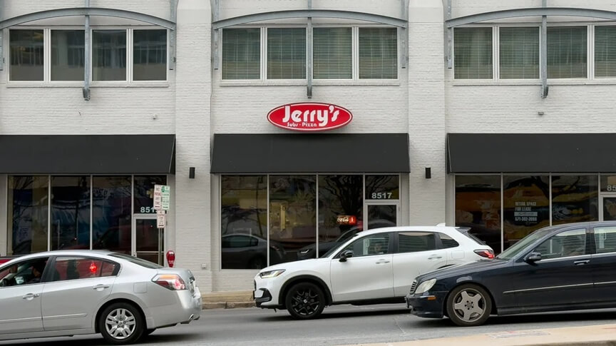 A Jerry’s Subs & Pizza restaurant located in a modern white brick building with large windows and black awnings. A small red oval sign displaying the restaurant’s name is mounted above the entrance. Several cars, including a white SUV and a black sedan, are parked along the busy city street in front of the restaurant.