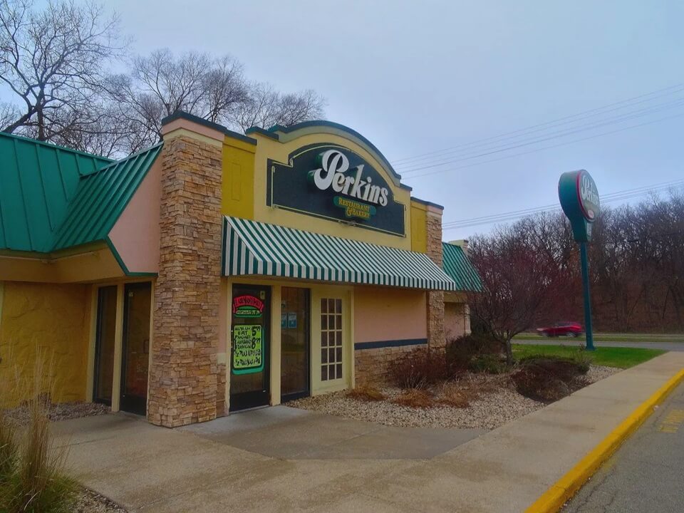 A Perkins Restaurant & Bakery location with a green metal roof, stone-accented walls, and a striped green-and-white awning over the entrance. The large Perkins sign is displayed on the building, with another tall sign standing near the road. The sidewalk leading to the entrance is lined with decorative rocks and shrubs, and a red car is seen driving in the background.
