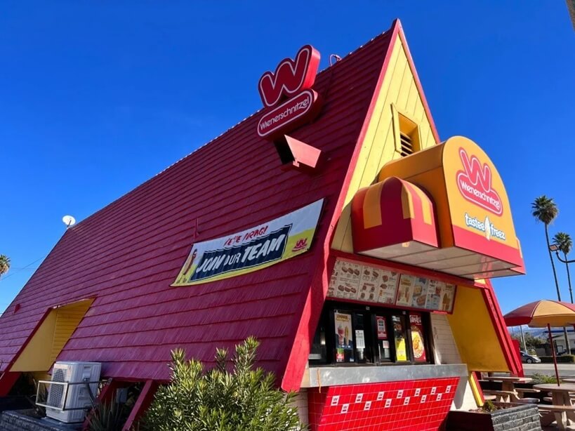 A classic Wienerschnitzel fast-food restaurant with an iconic A-frame red and yellow exterior. The restaurant's logo, a red "W" with "Wienerschnitzel" written underneath, is displayed on the roof and above the ordering window. A banner on the side advertises job openings, and the menu is visible at the walk-up order counter under a red and yellow striped awning.