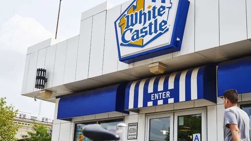A White Castle fast-food restaurant with a white and blue exterior, featuring the brand’s signature castle-like design. The large White Castle logo is prominently displayed above the entrance, which has a blue awning with white stripes and the word "ENTER" written on it. A young man is seen walking toward the entrance, while a train passes by in the background.