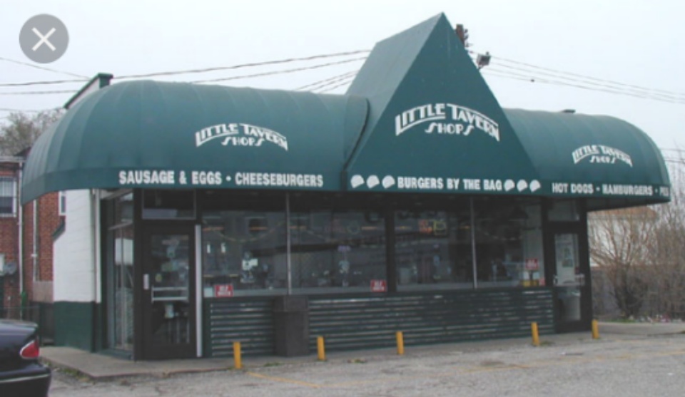 A vintage Little Tavern Shop restaurant with its signature green awning and pyramid-shaped roof. The awning displays the restaurant's name along with menu items like "Sausage & Eggs," "Cheeseburgers," "Burgers by the Bag," and "Hot Dogs." The small building has large glass windows, and the parking lot in front has a few yellow barriers.
