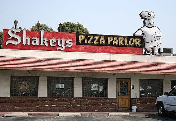 A classic Shakey’s Pizza Parlor restaurant with a retro red and black sign featuring the brand’s signature lettering. A large cartoon chef mascot wearing a hat with an "S" stands atop the roof, adding to the vintage charm. The building has a red-tiled awning, brick and cream-colored walls, and a wooden door with a "Closed" sign in the window.