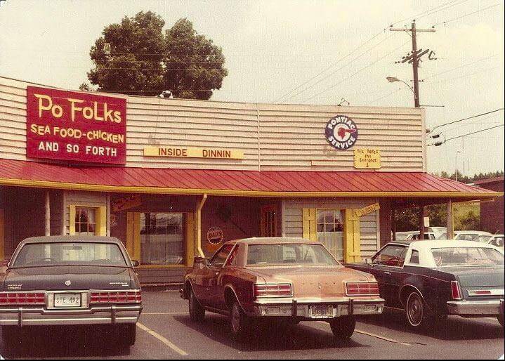A vintage Po Folks restaurant with a rustic wooden exterior, a red metal roof, and yellow shutters. A red sign on the left advertises "Po Folks SEA FOOD - CHICKEN AND SO FORTH," while a yellow sign above the entrance humorously reads "INSIDE DINNIN." Classic cars from the late 1970s or early 1980s are parked in front, enhancing the nostalgic atmosphere.