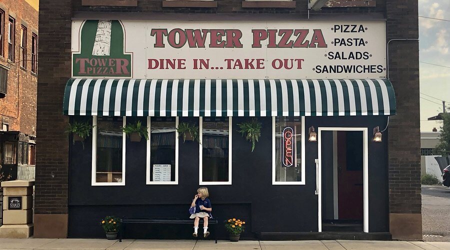 Tower Pizza, a small-town pizzeria housed in a brick building, features a vintage-style sign advertising pizza, pasta, salads, and sandwiches. A green and white striped awning shades the front windows, with a neon "OPEN" sign glowing near the entrance. A young girl sits on a bench outside, adding a charming, nostalgic feel to the scene.