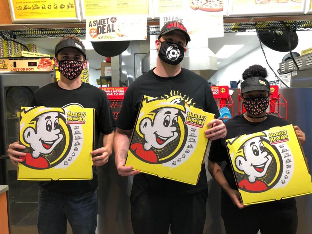 Three Hungry Howie’s employees wearing black uniforms, visors, and patterned face masks stand inside the pizzeria holding large yellow pizza boxes featuring the brand’s signature cartoon character. Behind them, menu boards, promotional posters, and stacks of pizza boxes are visible. The employees pose proudly, highlighting the restaurant’s recognizable branding and commitment to service.