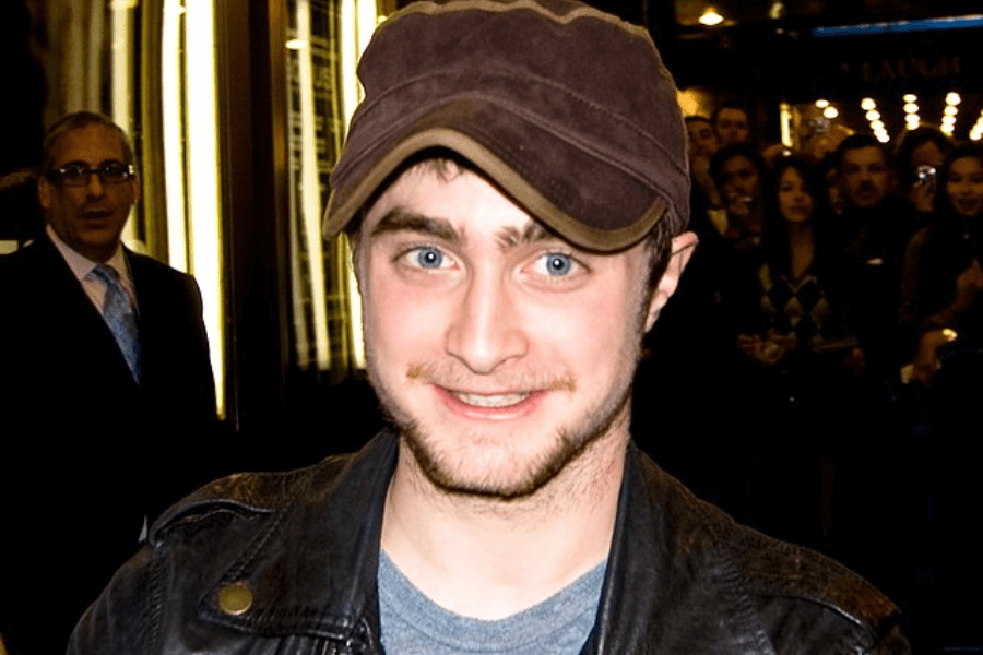 This image shows a person wearing a brown cap and a leather jacket, smiling while surrounded by a crowd of people in a lit urban environment, possibly at night.