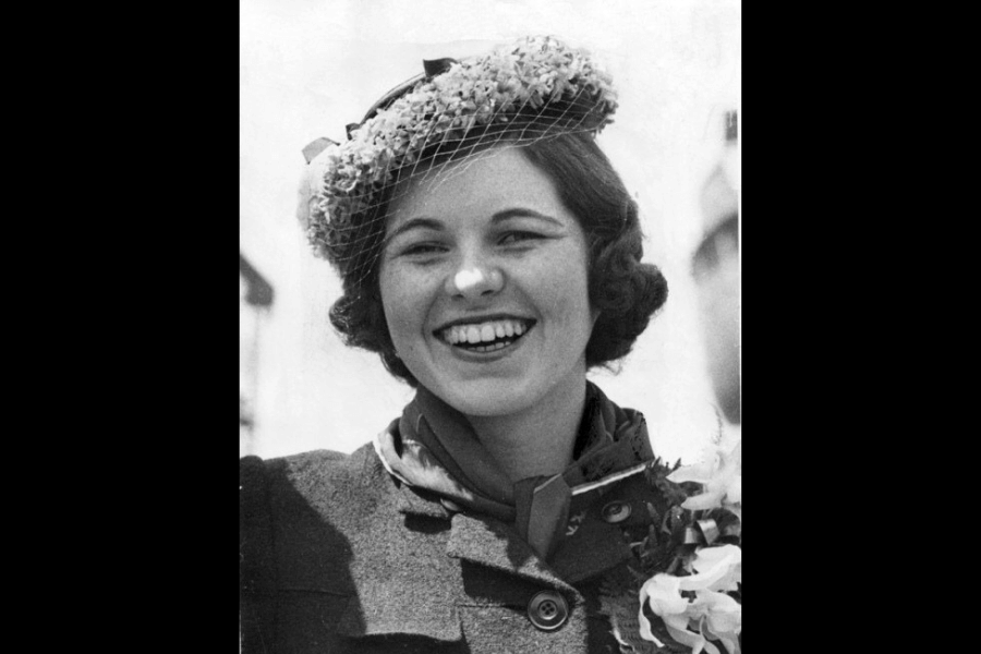 A black-and-white photograph of a smiling woman wearing a decorative floral hat with a small veil, paired with a scarf and a structured coat. Her expression exudes joy and confidence, capturing a vintage, timeless charm.