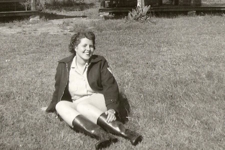 A black-and-white photograph of a woman sitting on grass outdoors, smiling warmly. She is dressed in a jacket, shorts, and knee-high boots, enjoying a relaxed moment in the sun with a rustic backdrop of wooden structures and greenery.