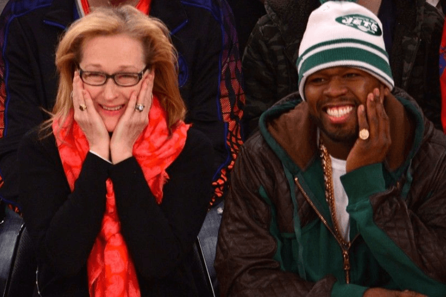 
This image shows a woman and a man sitting next to each other, both smiling widely with playful expressions, as if enjoying a fun or surprising moment. The woman, wearing glasses and a vibrant red scarf, has her hands on her cheeks, while the man, dressed in a green jacket and a white beanie with a sports logo, mirrors her pose with a big grin. The setting appears to be a lively event, likely a sports game, with a colorful crowd in the background.