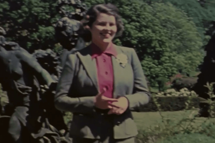 A color photograph of a woman standing outdoors in a lush garden with greenery and sculptures in the background. She is wearing a tailored suit with a red blouse underneath, smiling warmly while gesturing with her hands, exuding a poised and confident demeanor.