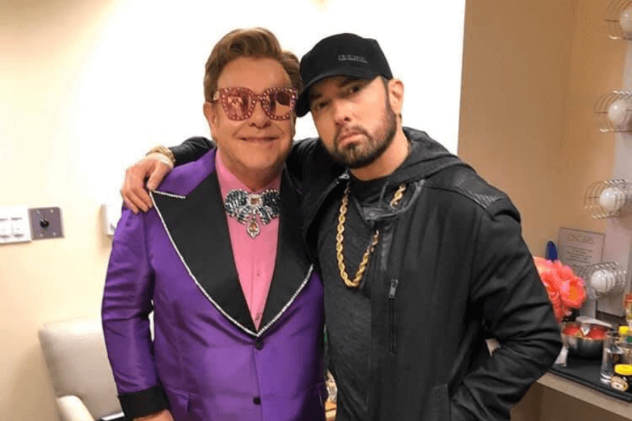 
This image shows two men posing together in what appears to be a backstage or dressing room setting. One wears a vibrant purple suit with a jeweled bow tie and oversized pink sunglasses, exuding a bold and flamboyant style, while the other sports a black hoodie, baseball cap, and a gold chain for a casual yet edgy look. The room's decor includes a makeup station and colorful flowers, adding to the behind-the-scenes atmosphere. 