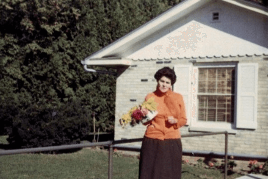 A color photograph of a woman standing in front of a small brick house with white shutters, holding a bouquet of colorful flowers. She is wearing an orange sweater and a dark skirt, smiling slightly, with lush greenery and a well-maintained lawn in the background.