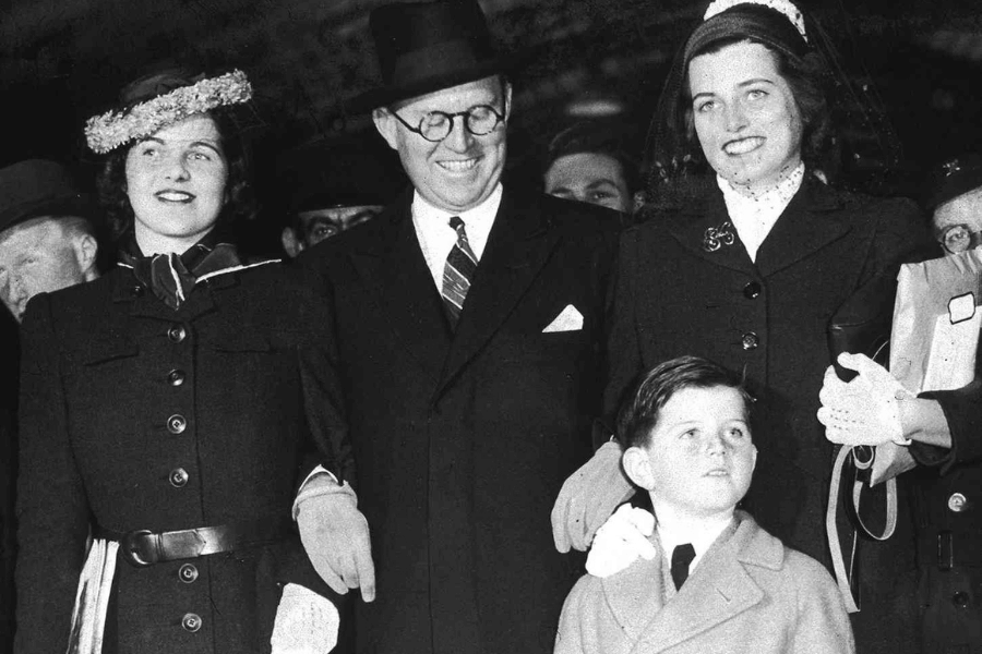 A black-and-white photograph of a family group, including two women, a man, and a young boy, all dressed in formal attire. One woman wears a floral hat and a buttoned coat, while the other has a veil and gloves. The man in the center wears a suit and tie with a hat, smiling warmly, as the boy in the front gazes upward, creating a moment of poised yet lively interaction.