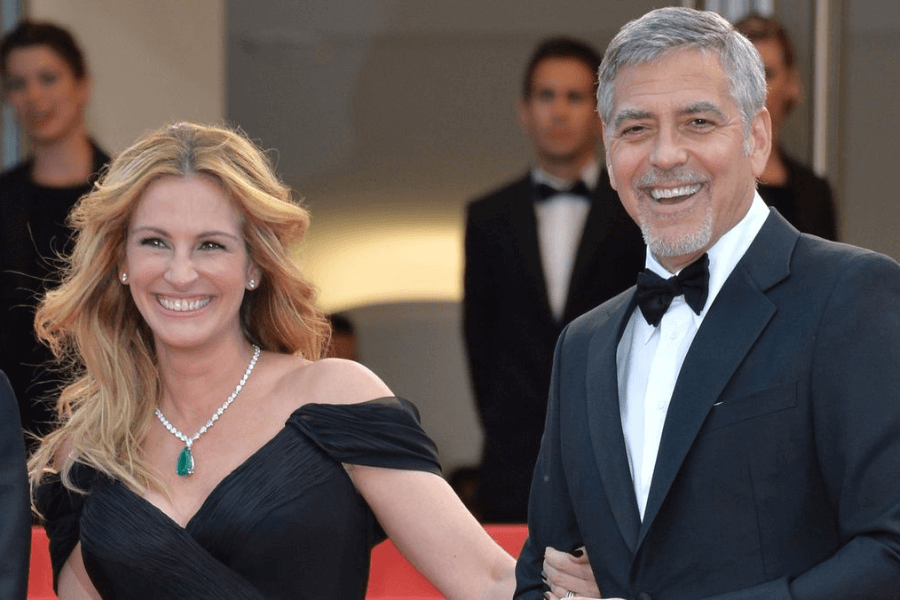 
This image shows a man and a woman dressed in formal attire, smiling warmly while walking arm-in-arm, likely at a red carpet event. The woman wears a stunning off-the-shoulder black gown paired with a statement emerald necklace, while the man looks dapper in a classic black tuxedo. The blurred background includes other attendees in formalwear, adding to the elegant and celebratory atmosphere. 