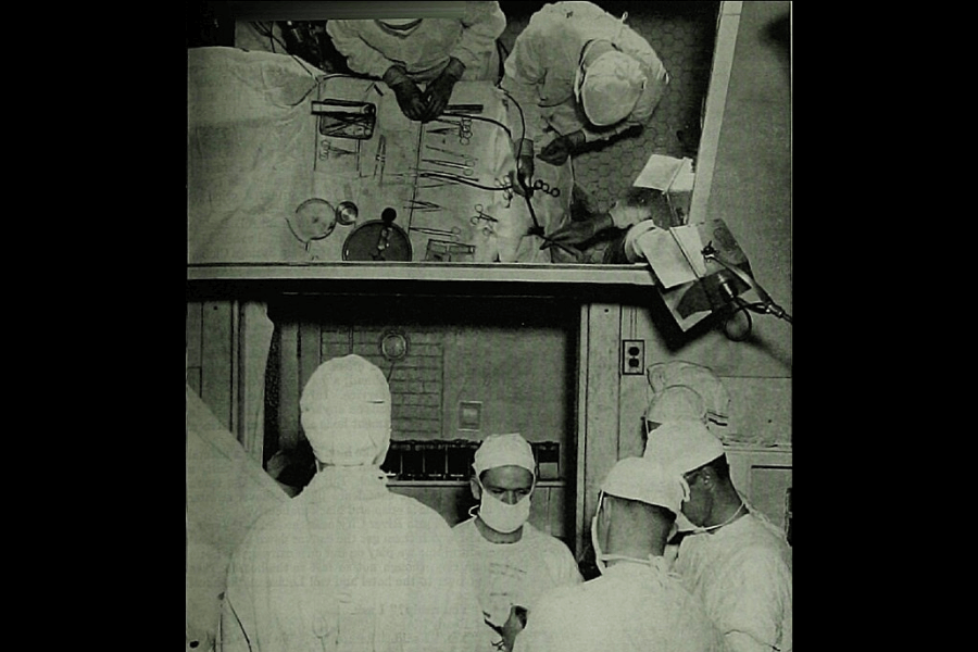 A black-and-white photograph depicting a surgical operation in progress. The scene includes surgeons dressed in scrubs, masks, and caps, surrounded by medical instruments and equipment. The image captures both a top-down view of the surgical tools and a ground-level perspective of the team working collaboratively in an operating room.