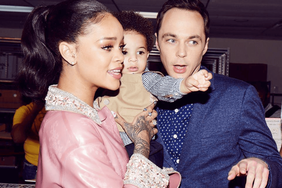 
This image captures a lively interaction between three people. A woman with her hair tied back, wearing a pink outfit adorned with sparkling details, holds a young child dressed in a beige vest and patterned shirt. A man in a blue blazer with a polka-dot shirt stands beside them, animatedly pointing, as the child also points in the same direction. The setting appears to be a casual indoor event with shelves and other people visible in the background. 