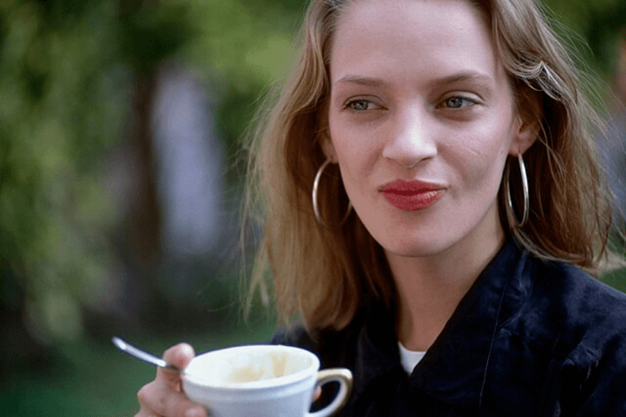 This image features a person with shoulder-length blonde hair, wearing hoop earrings and a dark outfit, holding a white teacup with a spoon inside. The outdoor setting includes a blurred green background, suggesting a relaxed moment in a garden or park.